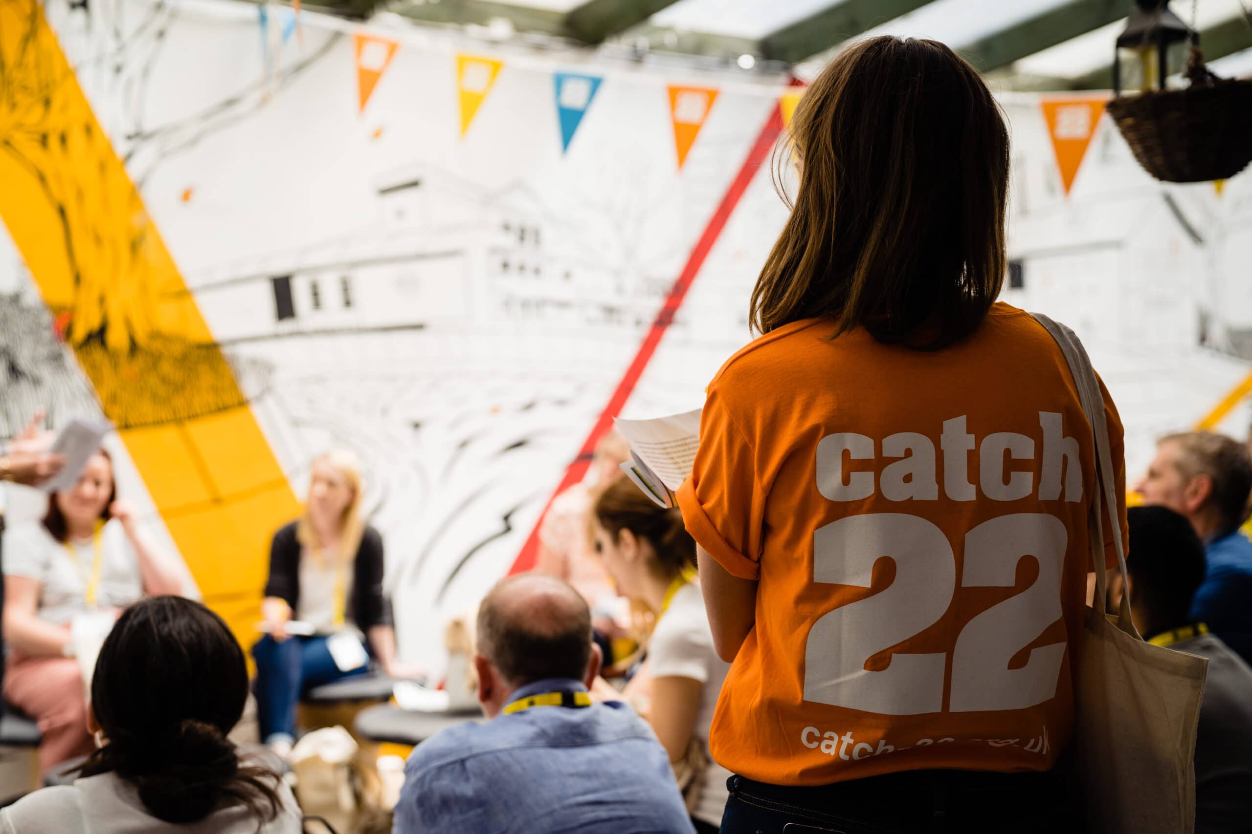 A woman stands in the foreground wearing an orange Catch22 tshirt with a Catch22-branded fabric tote bag. In the background, people are taking part in a workshop.
