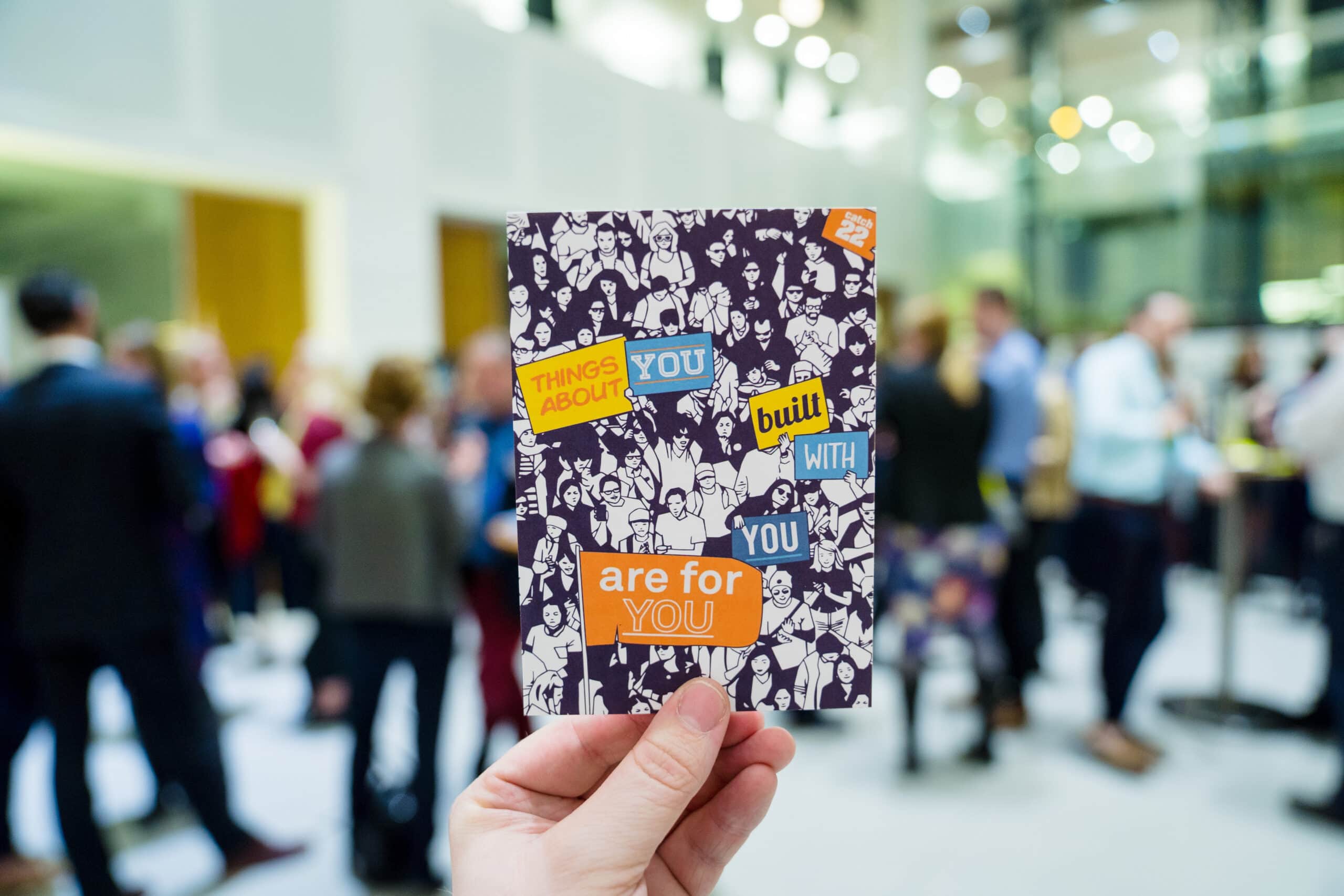 A hand holds out a Catch22-branded postcard which reads "Things about you, built with you, are for you". In the background, there are blurred people networking at an event.
