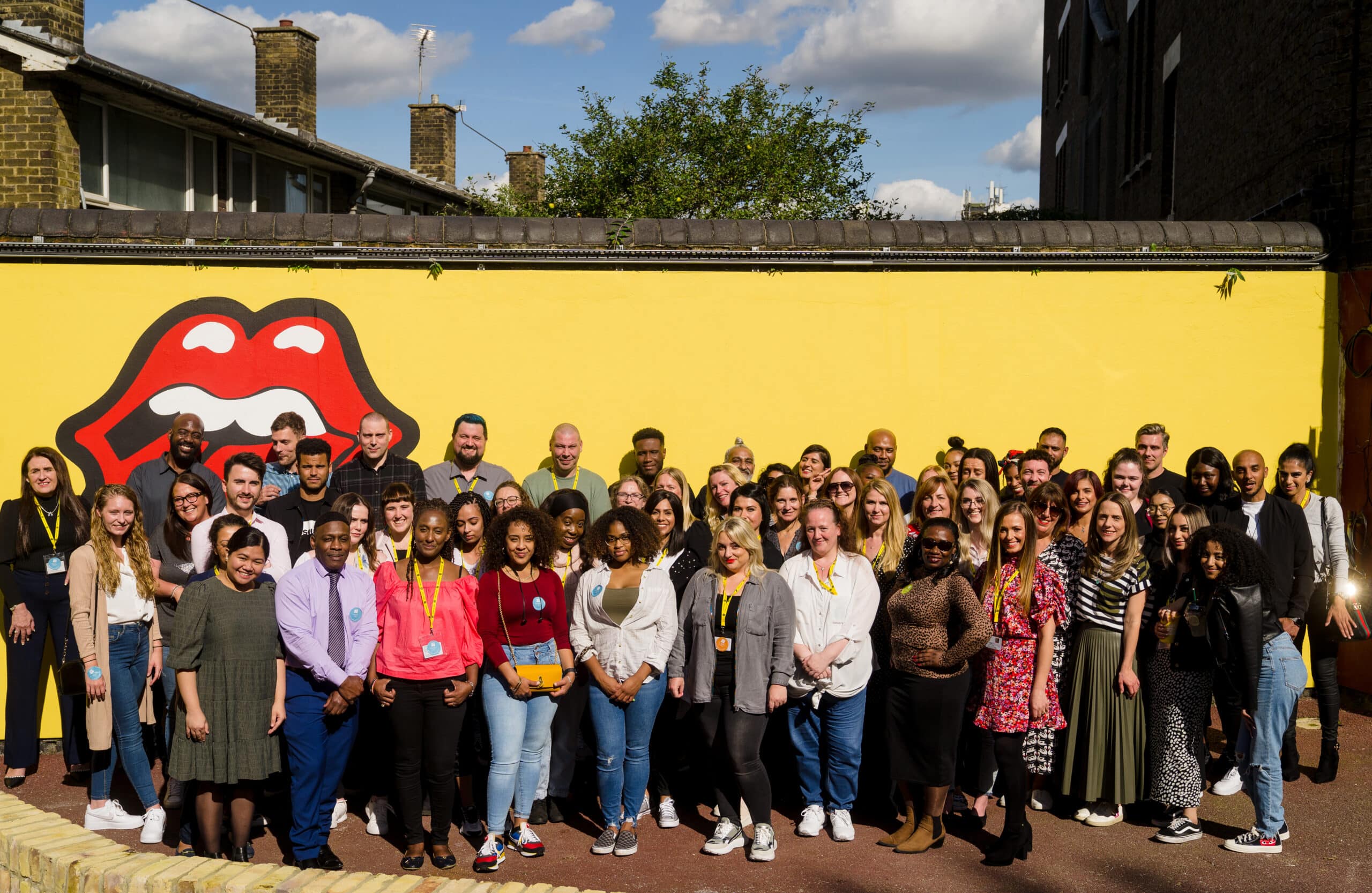 A large group of people from the Catch22 employability team stand in the sunshine in front of a yellow wall.