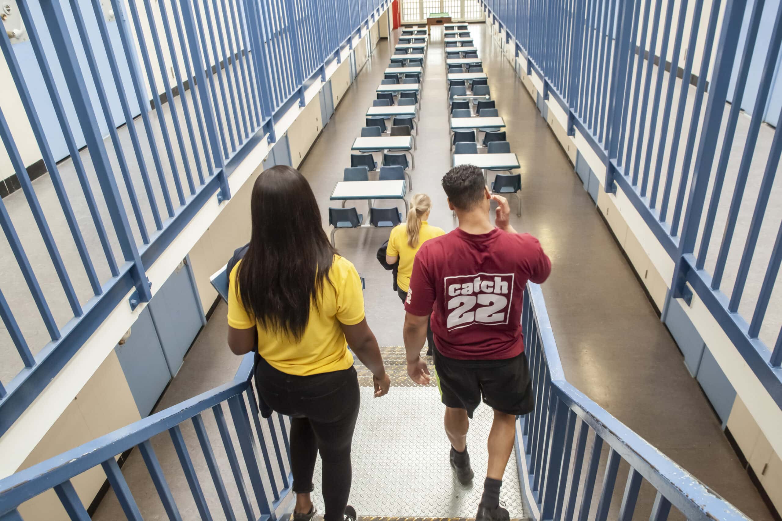 A man and two women walk down the stairs at HMP Thameside wearing Catch22 uniform. At the bottom of the stairs, there are rows of tables and chairs.