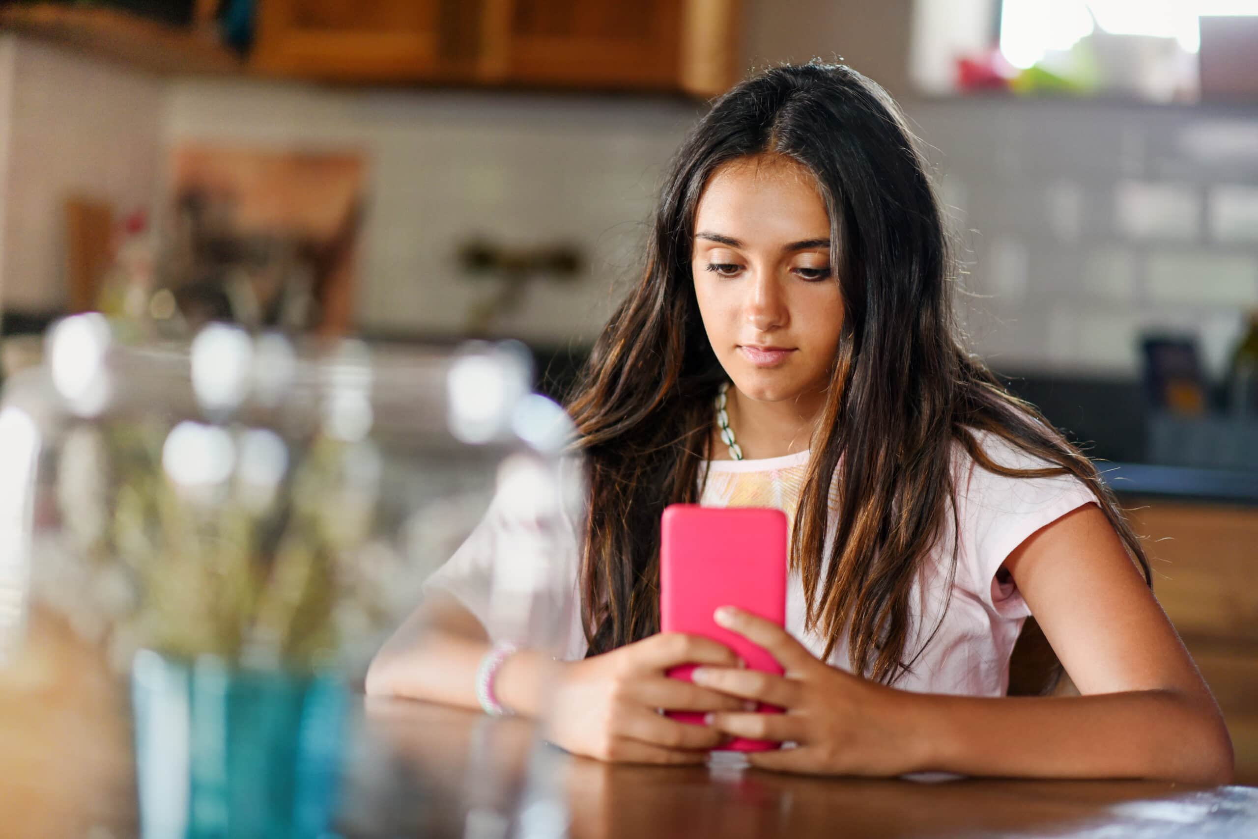 A teenage girl sits at the dining table reading messages or browsing the internet on her phone.