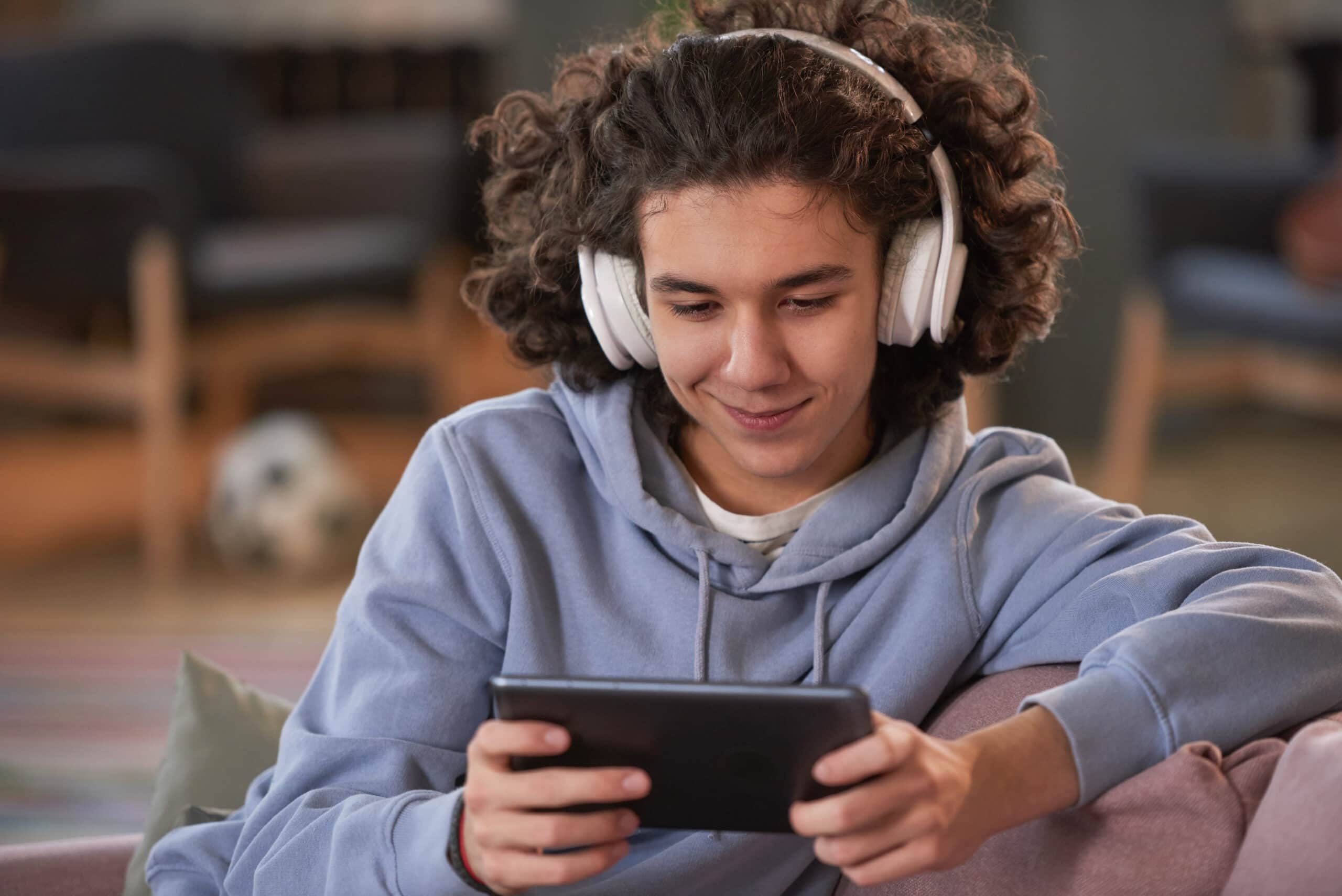 A teenage boy sits on the sofa watching something on his phone whilst listening to it using wireless headphones.