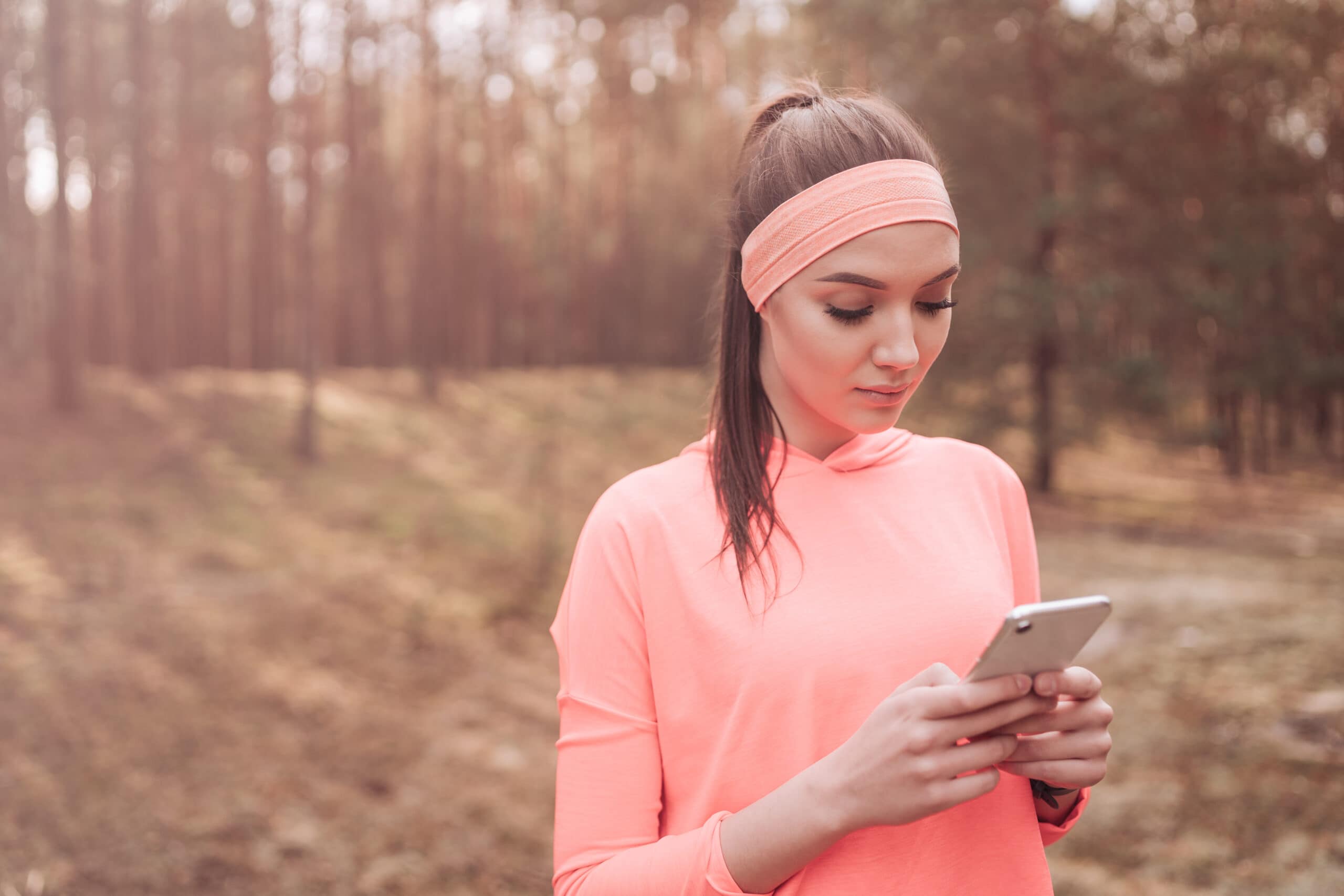 A young woman with a ponytail checks her phone whilst walking through a forest.