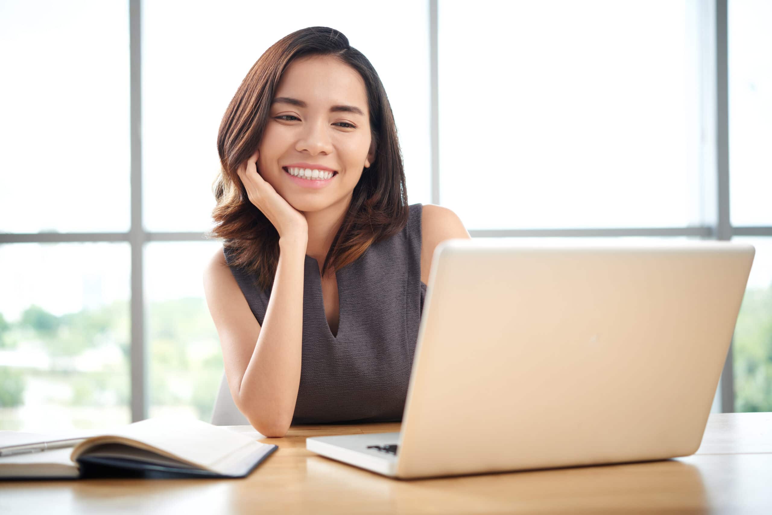 A young woman, facing the camera, sits in front of a laptop smiling. To her side is an open notebook.