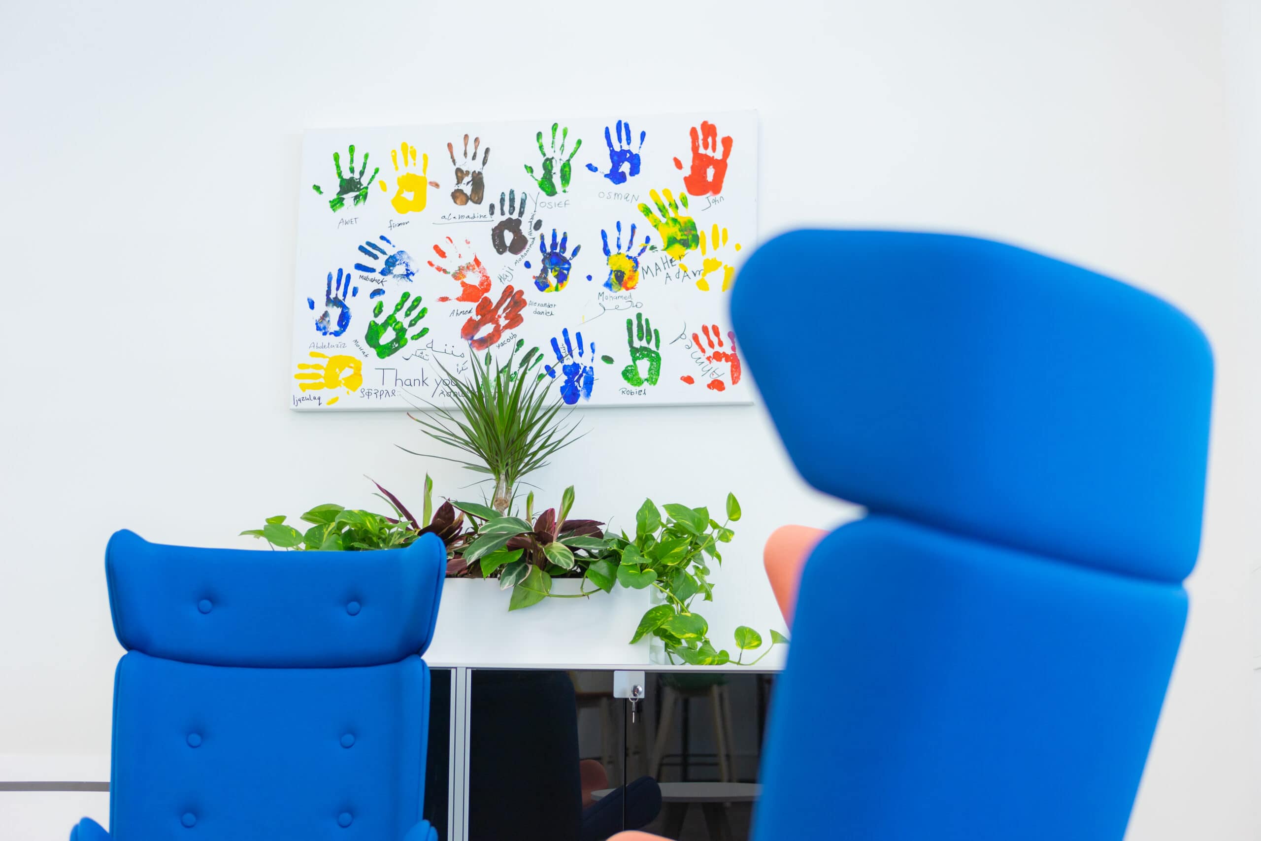 Two blue chairs are positioned in the background in front of a mural with colourful painted hand prints signed by the individuals who made them.