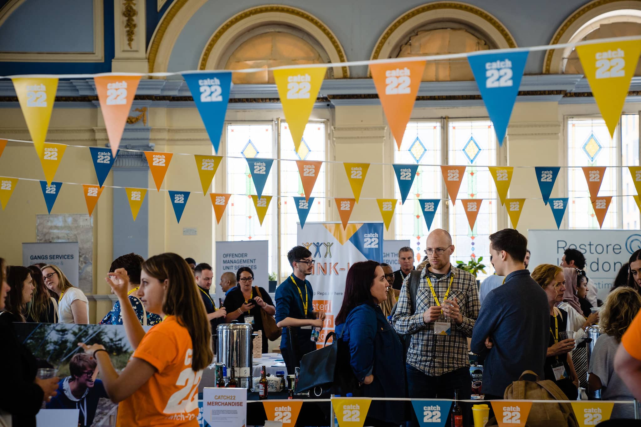 People gather around stands at an event, networking with their colleagues. Above them is Catch22-branded bunting.