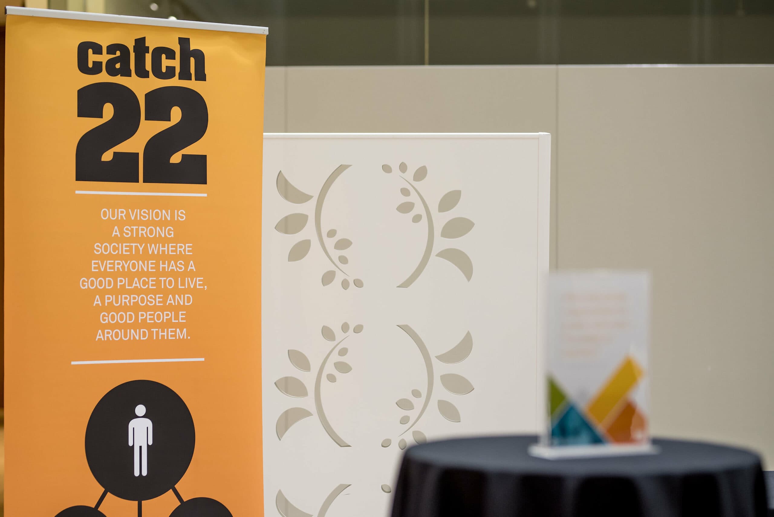 A yellow roll-up banner is set up at an event which reads "Catch22: Our vision is a strong society where everyone has a good place to live, a purpose, and good people around them."