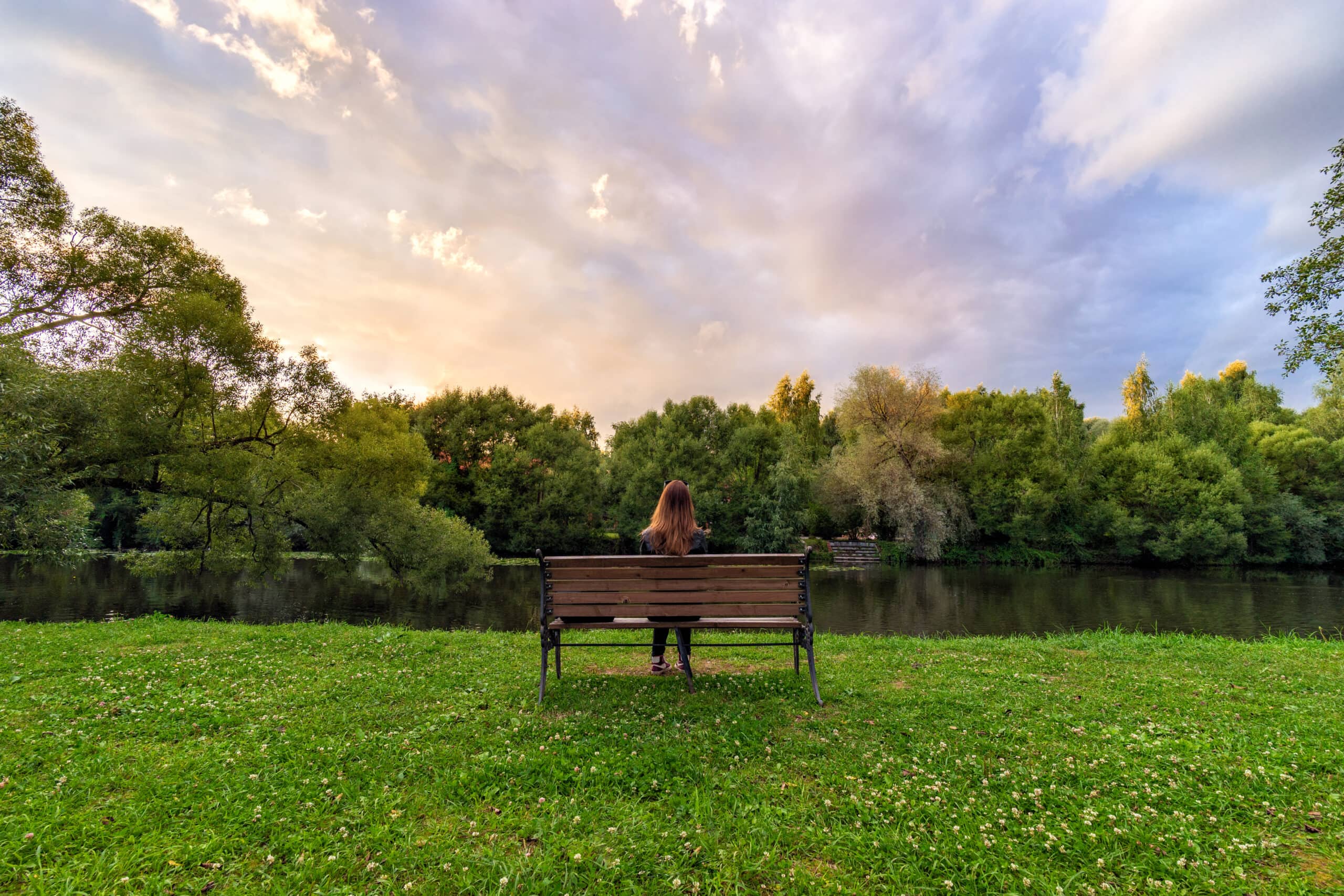 A young woman sits alone on a bench in the park as the sun sets.