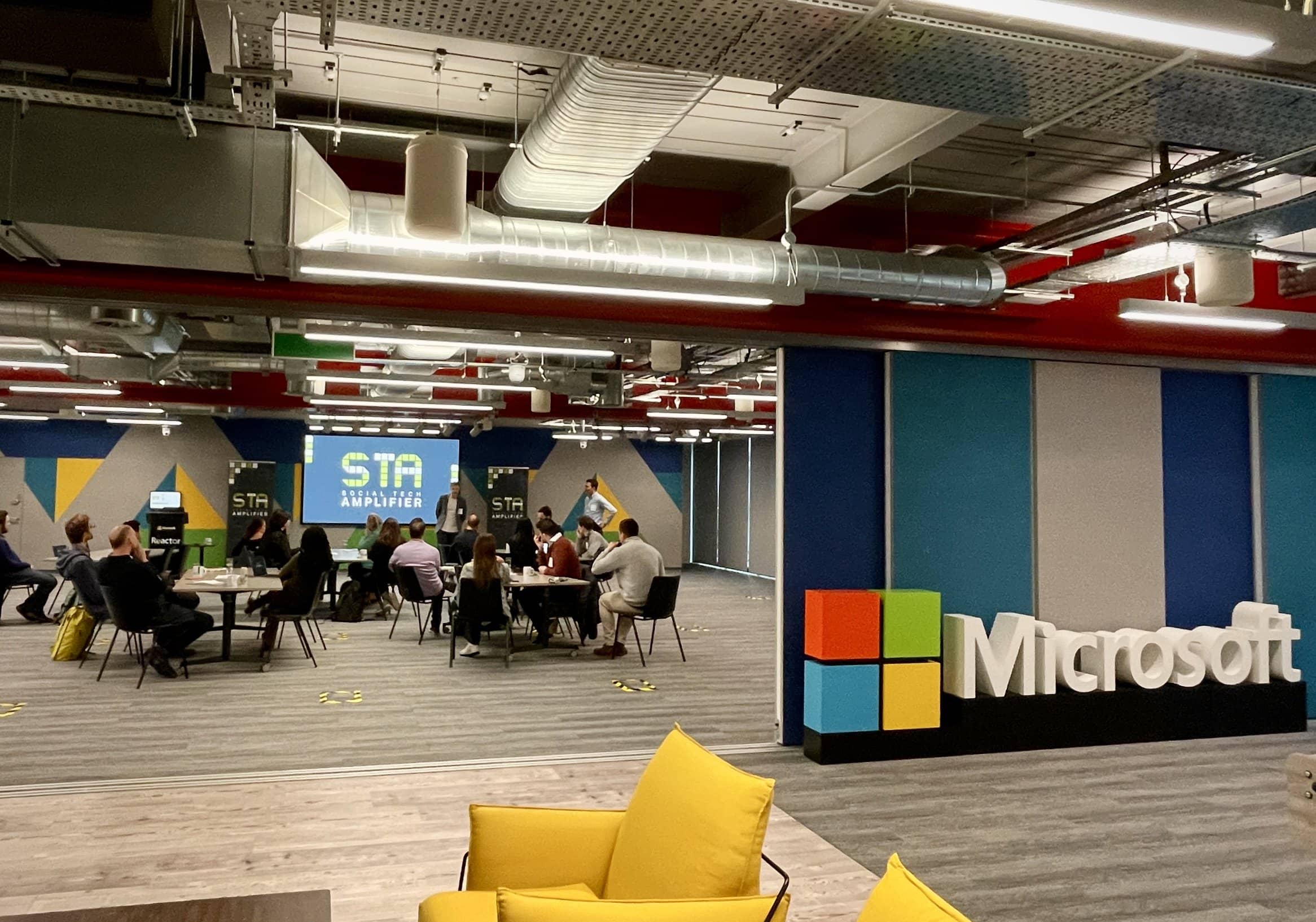 Taken through a doorway, professionals sit around tables listening to a speaker who stands in front of a presentation screen which shows the Social Tech Amplifier logo on it. In the foreground, the Microsoft logo can be seen.