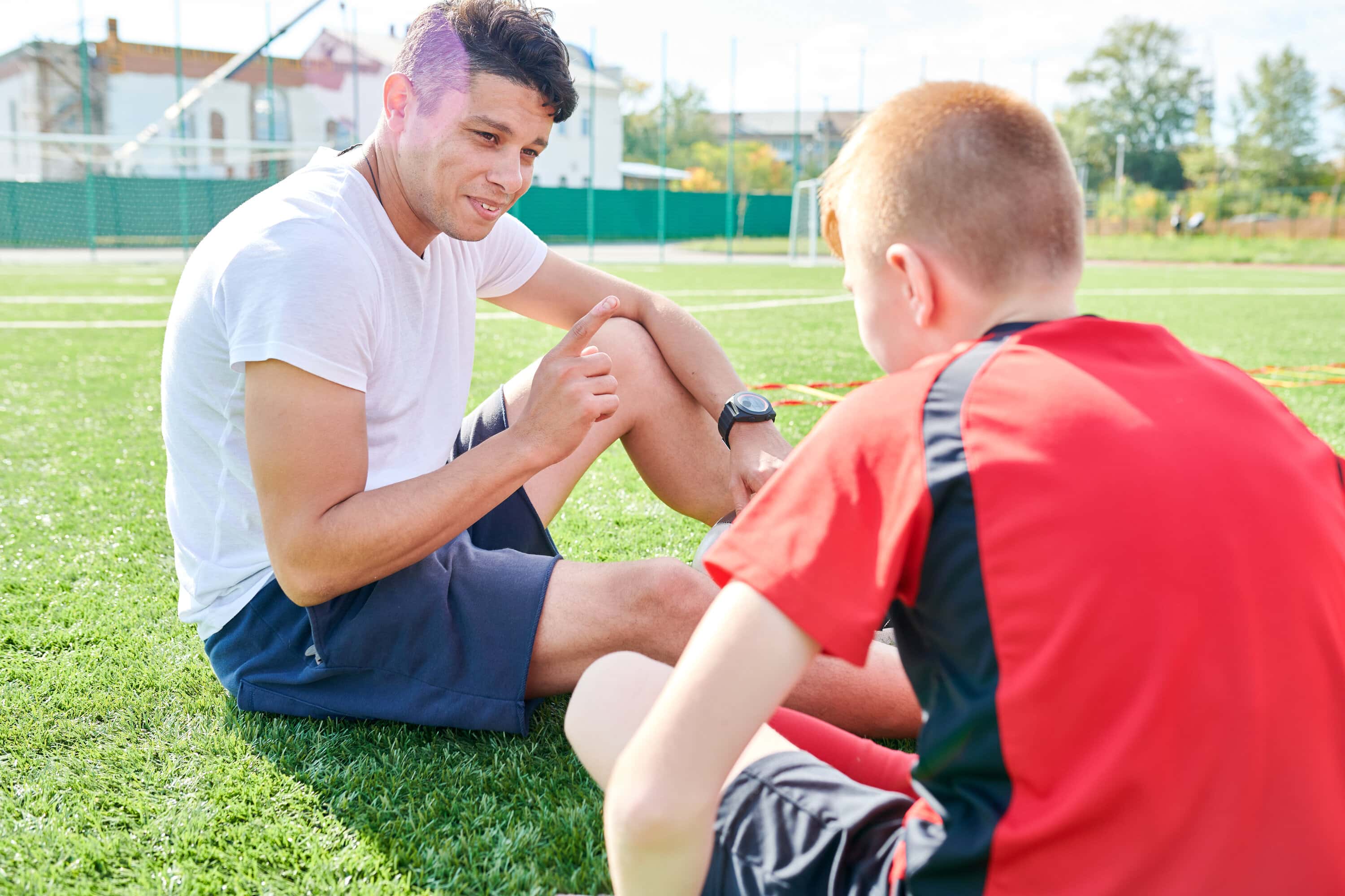 A male football coach talks to a teenage football player whilst sitting on the grass outside.