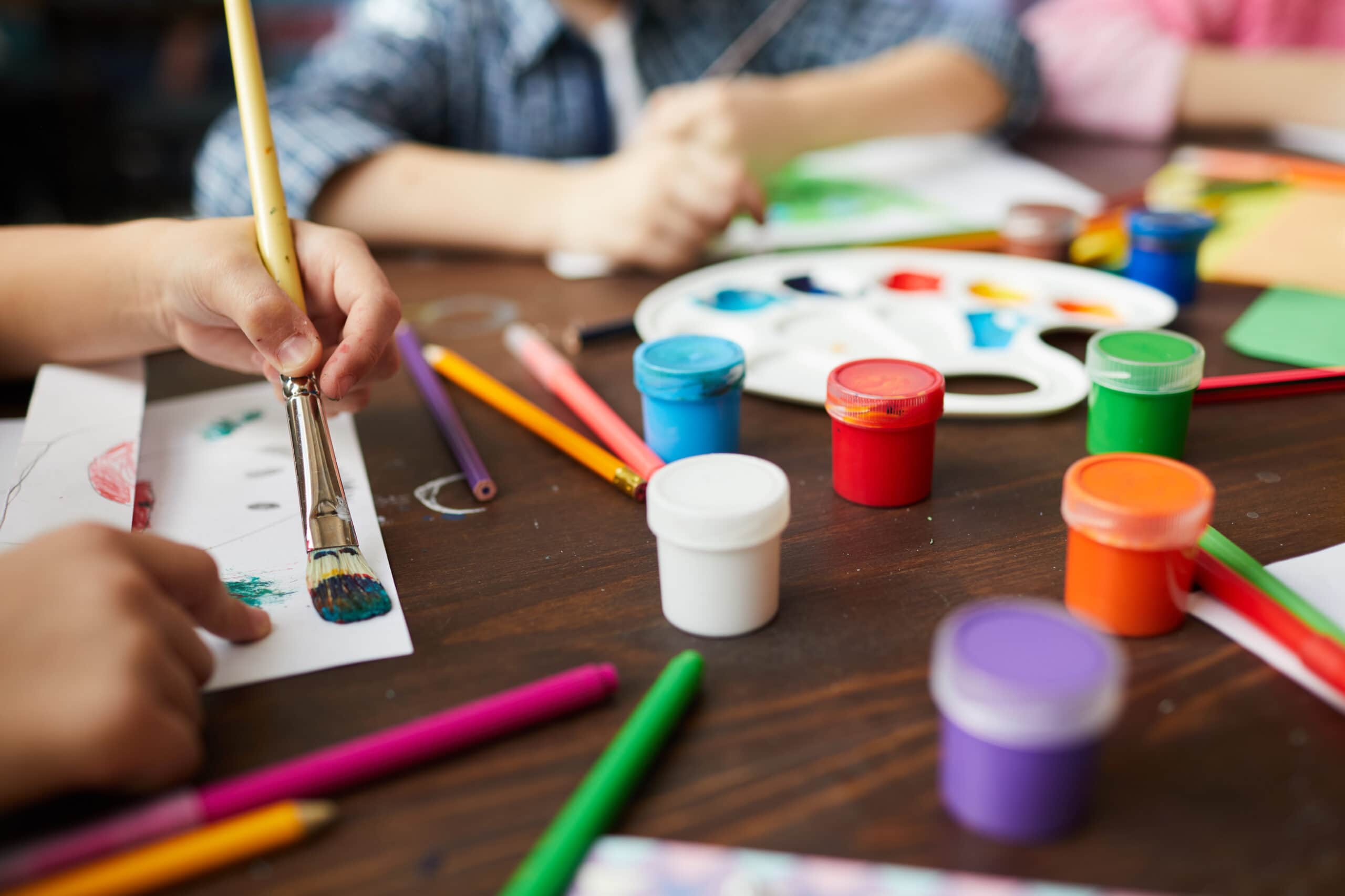 Close-up of children painting pictures, focus on art supplies paints, pencils and crayons.