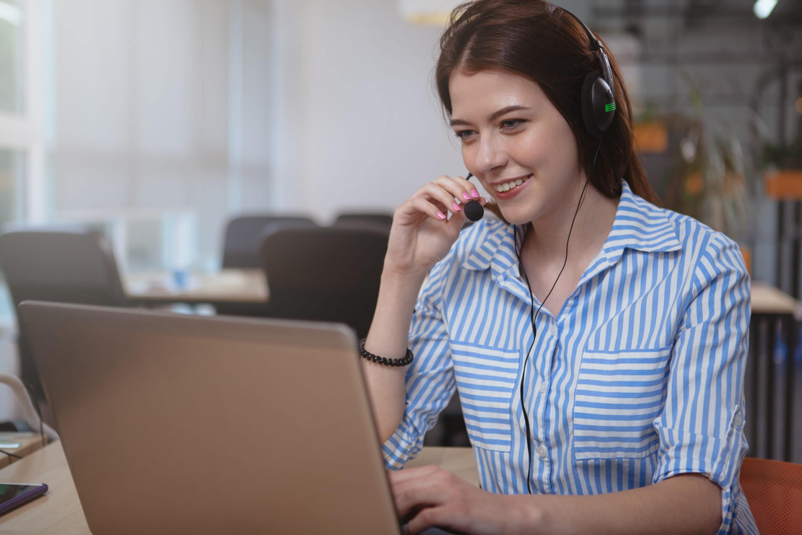 A female call centre worker, wearing a headset, answers a call whilst sitting in front of a laptop.