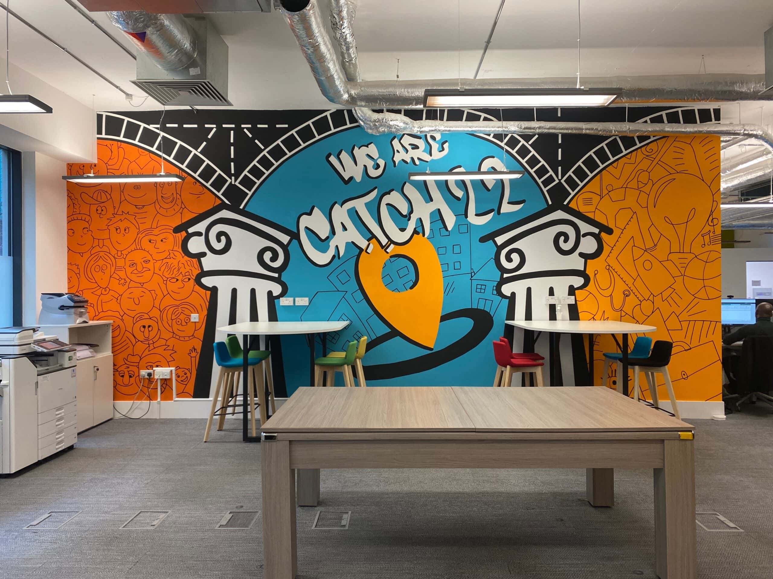 A bright coloured mural is painted on the wall of an office, which reads "We are Catch22". The mural depicts Catch22's 3Ps - people, place and purpose. In front of the mural, you can see office equipment including printers, desks and chairs, and a pool table.