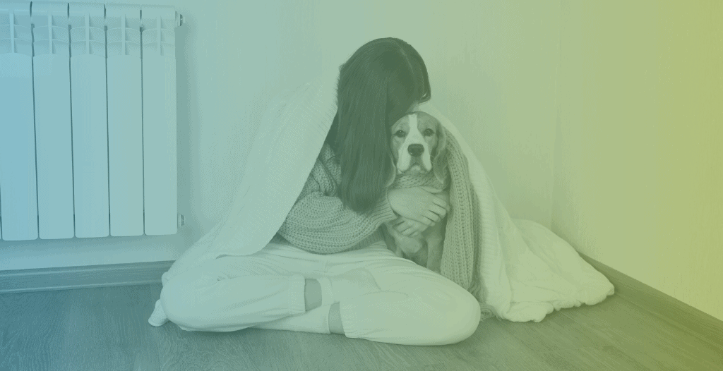 A young woman sits on the floor covered with a blanket. She hugs a dog and her face is covered by her hair as she does so.