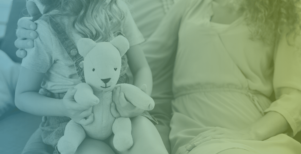 A child sits on an adult's lap, next to another adult who has an arm around their shoulder. She holds a teddy bear on her lap. None of their faces can be seen.