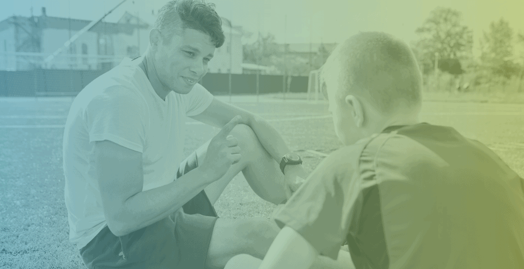 A male football coach talks to a teenage football player whilst sitting on the grass outside.