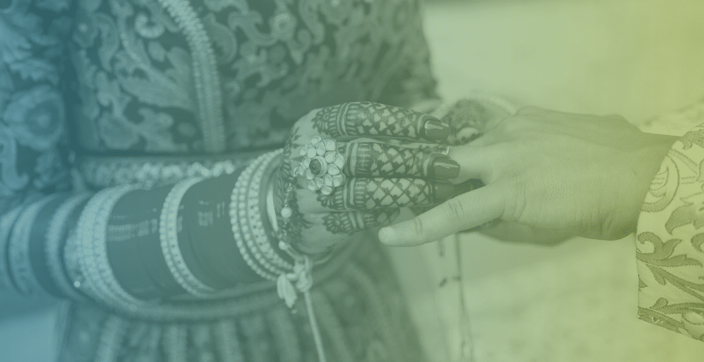 Close-up of a woman in a traditional wedding outfit placing a ring on the hand of a man. She has a lot of bracelets on her arms and her hands have mehndi designs applied to them.