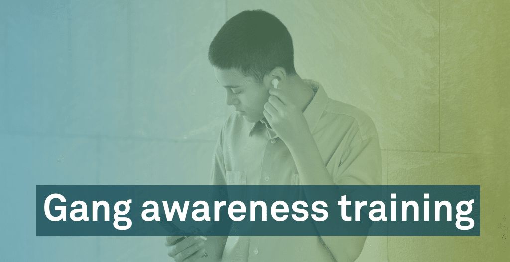 A young man stands in front of a wall. He places a wireless earphone into one ear. He is looking at his phone in his other hand. Overlaid is text that reads "Gang awareness training".