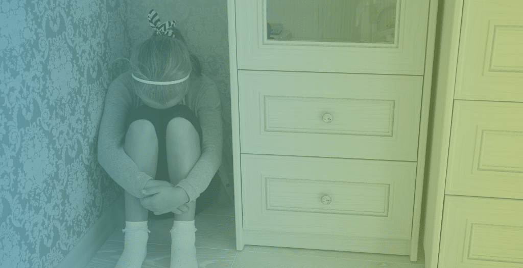 A young girl sits on the floor in a small space next to a cupboard. She rests her head on her knees and wraps her arms around her legs which are drawn up to her chest.