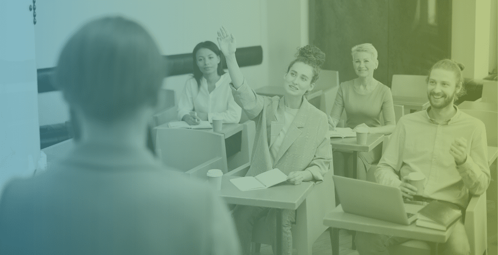 Adults sit at classroom desks making notes whilst listening to a presentation. A woman raises her hand to ask a question. The back of the teacher's head can be seen in the foreground.