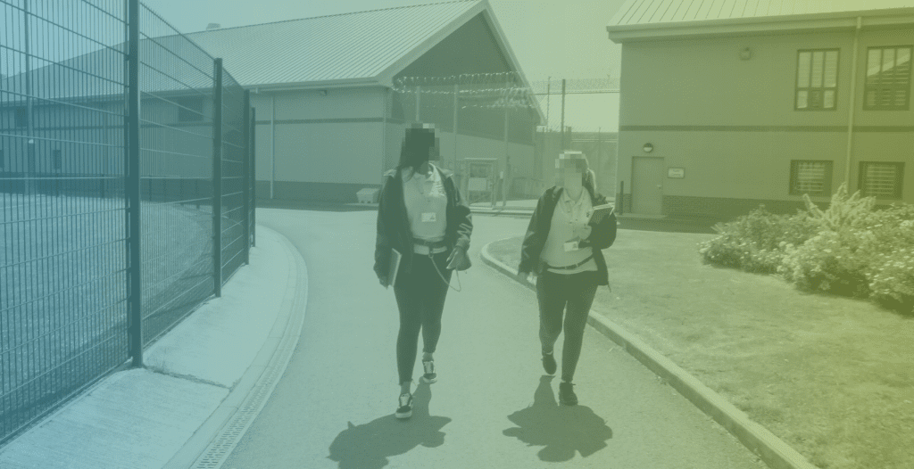 Two prison workers walk through the grounds of HMP Thameside. Both carry notebooks. The prison can be seen in the background.