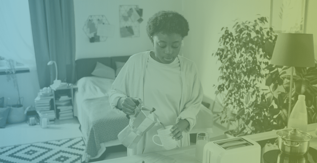 A young woman makes herself coffee at home. A studio flat can be seen in the background.