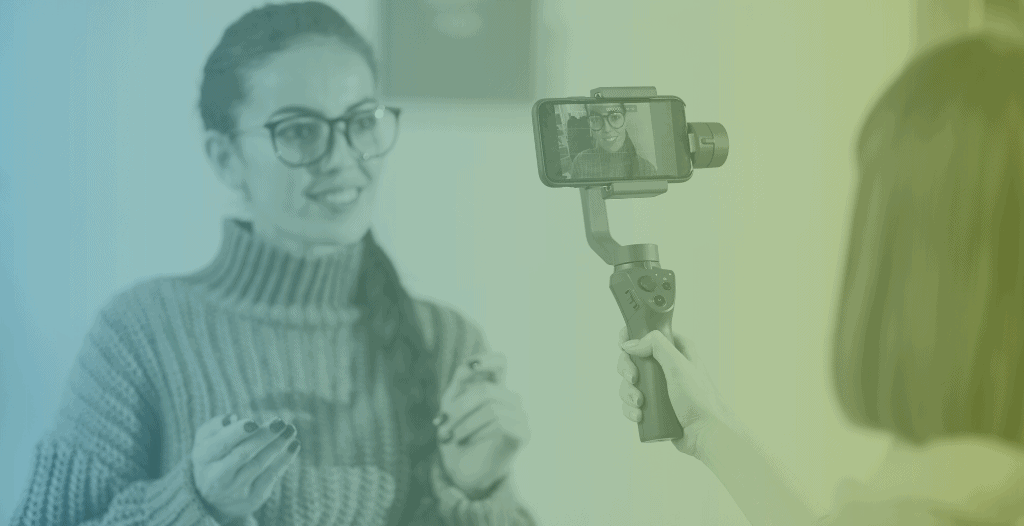 A young woman films content using her phone. Her phone is held on a stabiliser by a friend.