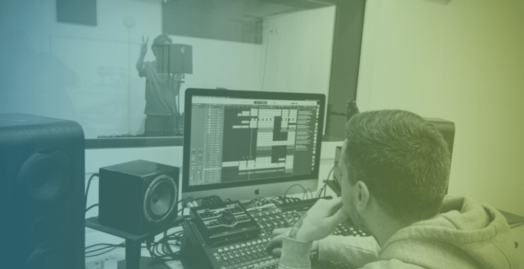A man operates recording studio software, whilst a young person records audio within a sound booth.