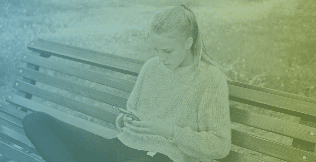 A teenage girl wearing casual clothes sits on a bench looking at her phone.