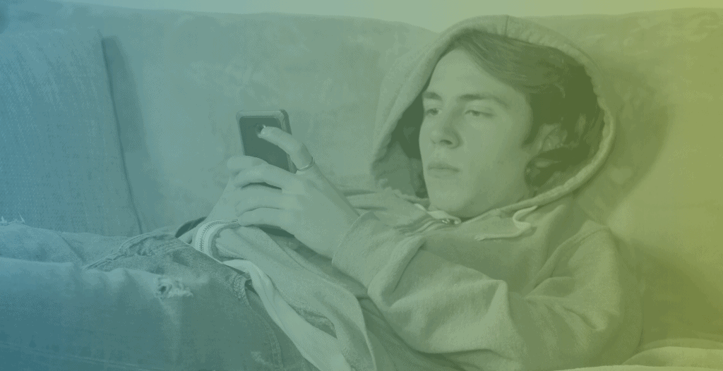 A teenage boy browses his phone whilst laying on the sofa. He is wearing a hoodie with the hood up.