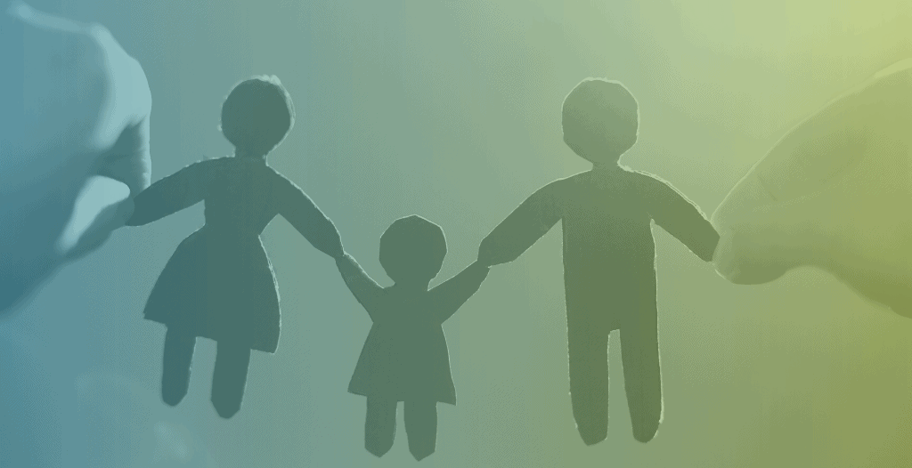 Paper cut-outs of three people - two adults and a child - are held up to the sky. The three paper figures are holding hands.