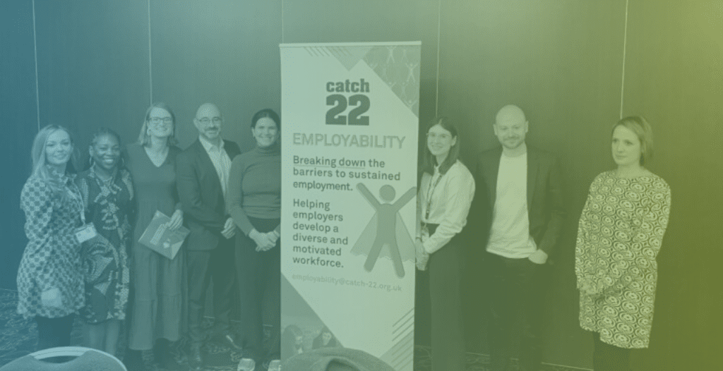 Catch22 staff smile at the camera, stood next to a roll-up banner which reads "Catch22 Employability: Breaking down the barriers to sustained employment. Helping employers develop a diverse and motivated workforce."