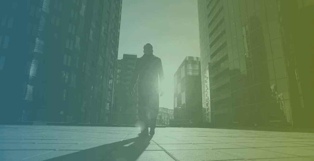 A man walks through a city, with tall buildings around him. The sun shines through the buildings behind him.