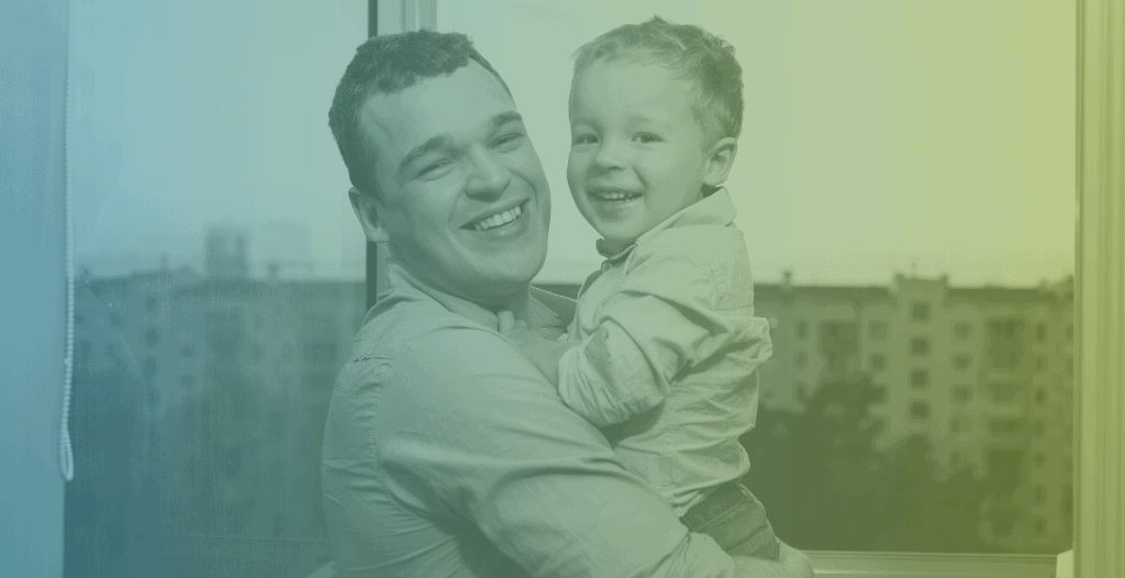 A man holds his young son in front of a large window. Both are smiling at the camera.