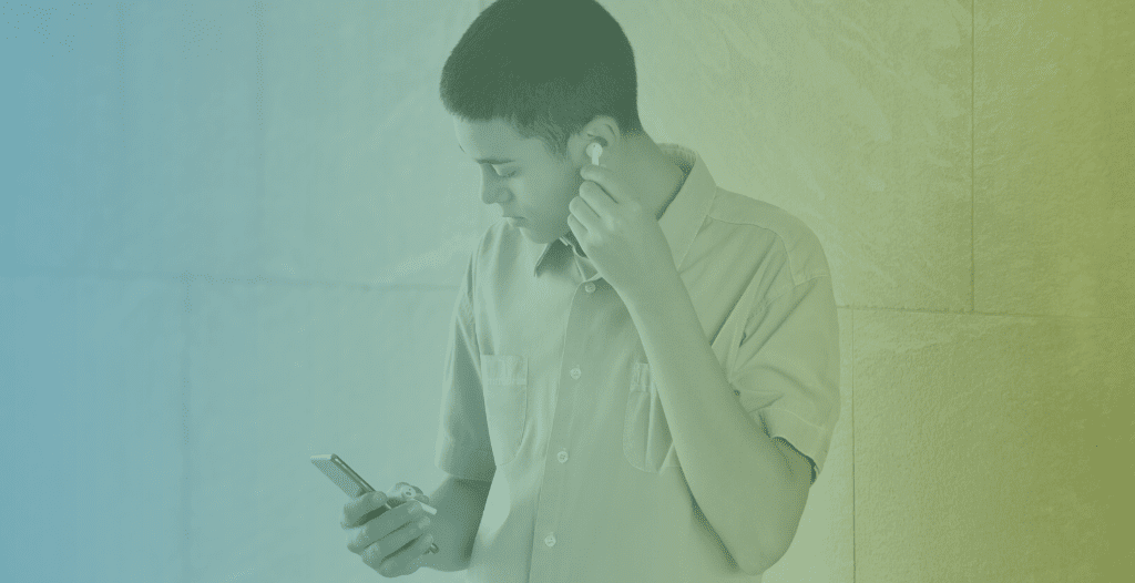 Young man listening to music on a mobile phone adjusting the earbud in his ear for better sound against a white stone commercial building wall.