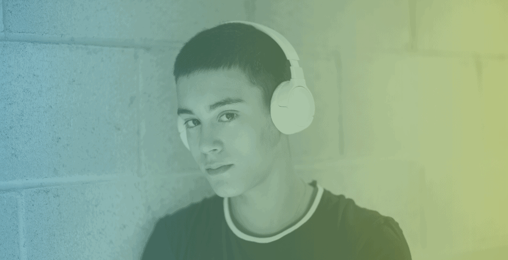 A teenage boy stands against a brick wall wearing over-the-head headphones. He looks at the camera from the side.