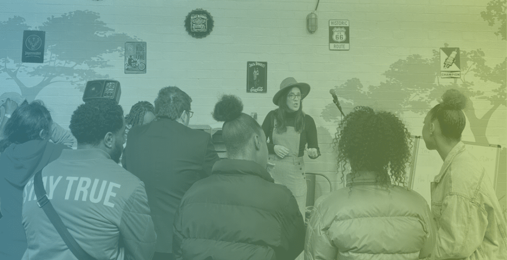 A woman wearing a hat presents to a group of young creatives who would like to start a career in a creative industry. The photograph is taken from the back of the crowd.