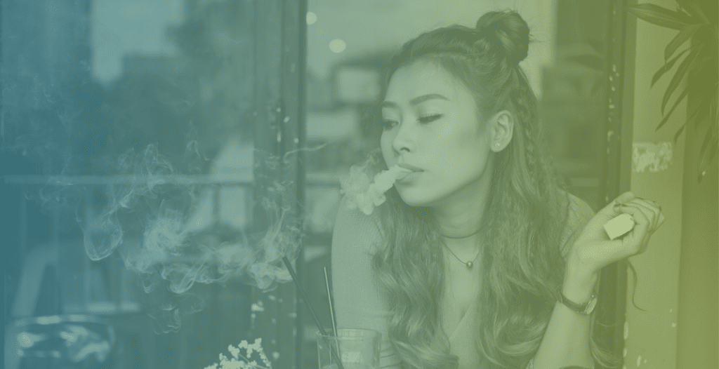 A young woman sits at a cafe using a vape. She breathes out the vapour into the air.
