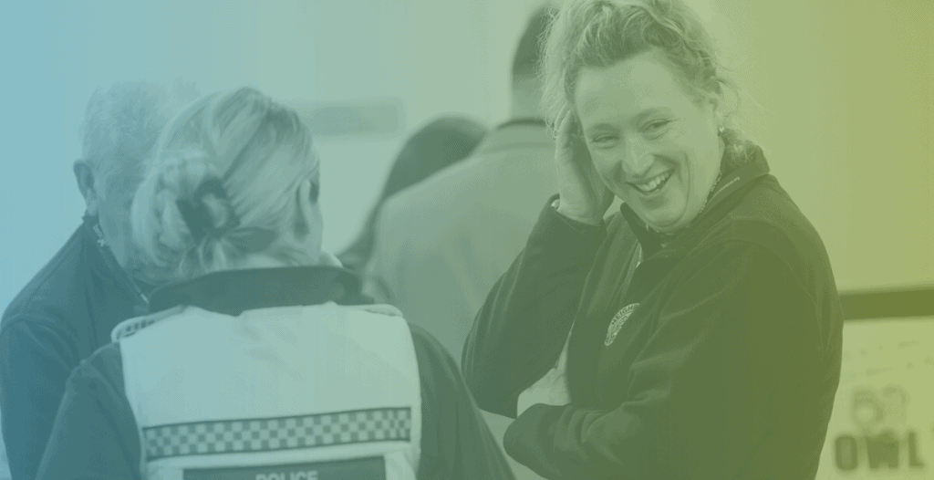 Two women laugh whilst having a conversation. One has her back to the camera but is wearing a police vest. The other is turned to look back towards the camera.